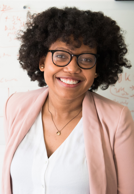 Woman standing and smiling in front of white board with academic notes written accross the board.