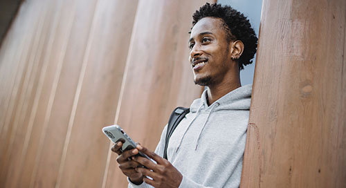 Male student walking out of a university building smiling with his phone in his hands