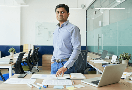 Man standing in a confereance room, he is smiling and wearing a blue shirt and jeans.