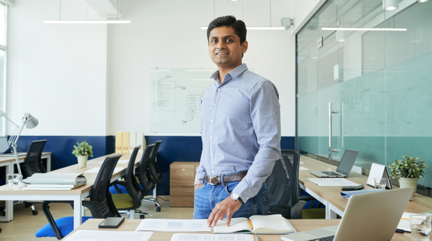 Man standing in a confereance room he is smiling and wearing a blue shirt and jeans.