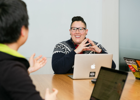 People sitting on a table with their laptops. Main person in focus is smiling, wearing glasses, a sweater and has their hands touching over their computer. 